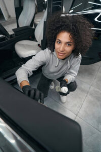 Fine Shine Mobile Wash A Fine Shine Mobile Wash expert, wearing gloves, cleans the interior of a car with a spray bottle while kneeling by the open door on a tiled floor.
